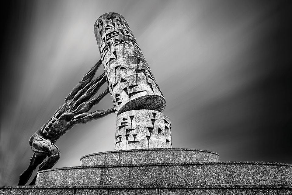 A black and white photo of a human form with many arms but no head pushing a column adorned in cuneiform back on to its pedestal.