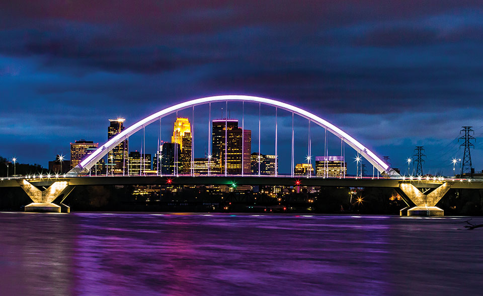 The skyline of Minnepolis seen through an arched bridge suspended over purple water