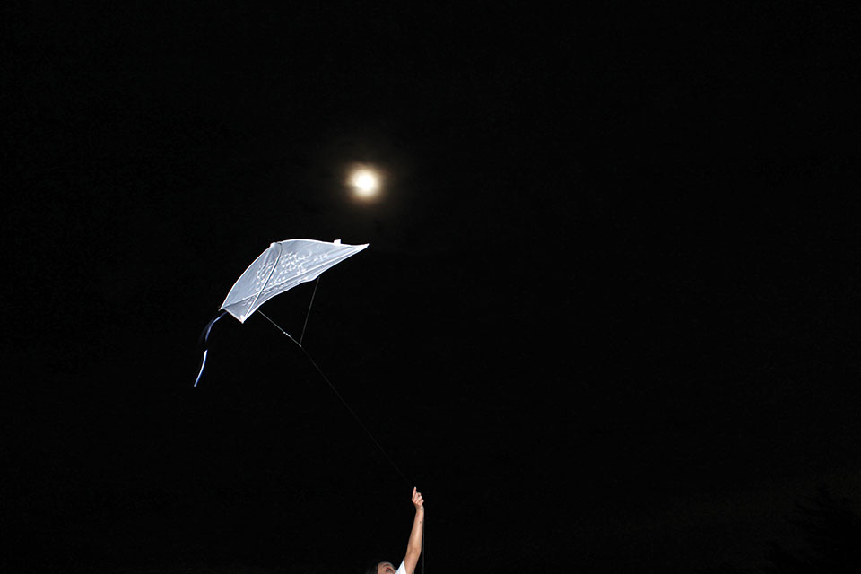 A photograph of a white kite held by an arm that stretches into the bottom of the frame with the moon hovering just above the kite