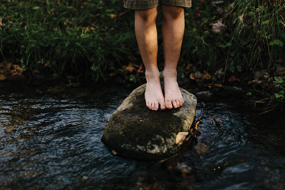 A pair of childish legs stand perched on a mossy rock in a brook