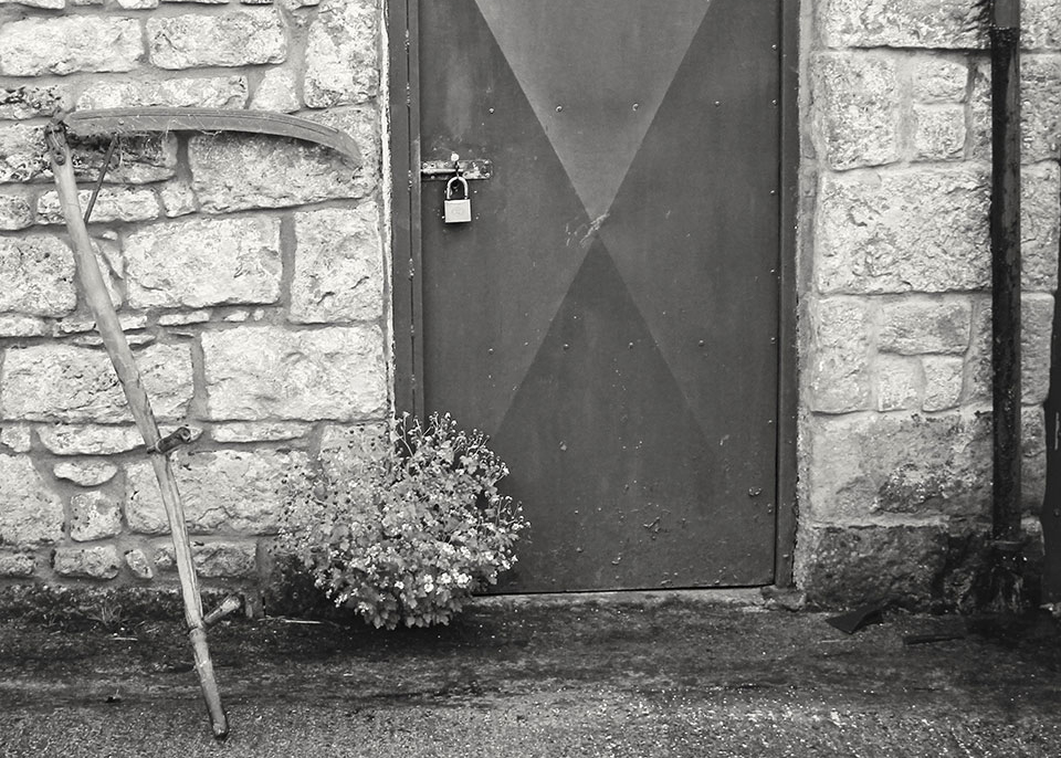 Black and white photograph of a rustic building with a padlock on the door. A scythe leans against the wall just to the left of the door