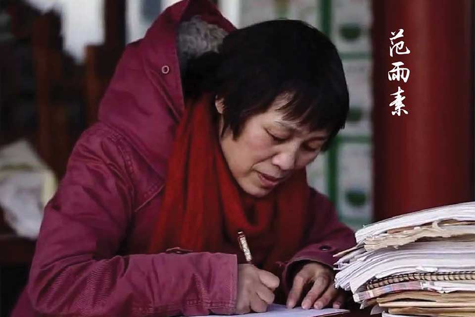 A photograph of a woman in a heavy red parka, hunched over a desk, writing