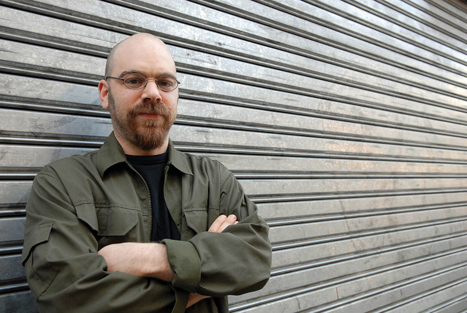 A photograph of Ariel Magnus, who stands with his arms crossed, in front of a lowered eaves