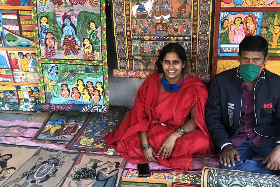 A woman clad in red sits with her husband (who is masked) on the ground. On the ground in front of them and mounted behind them on a wall of brightly colored paintings.