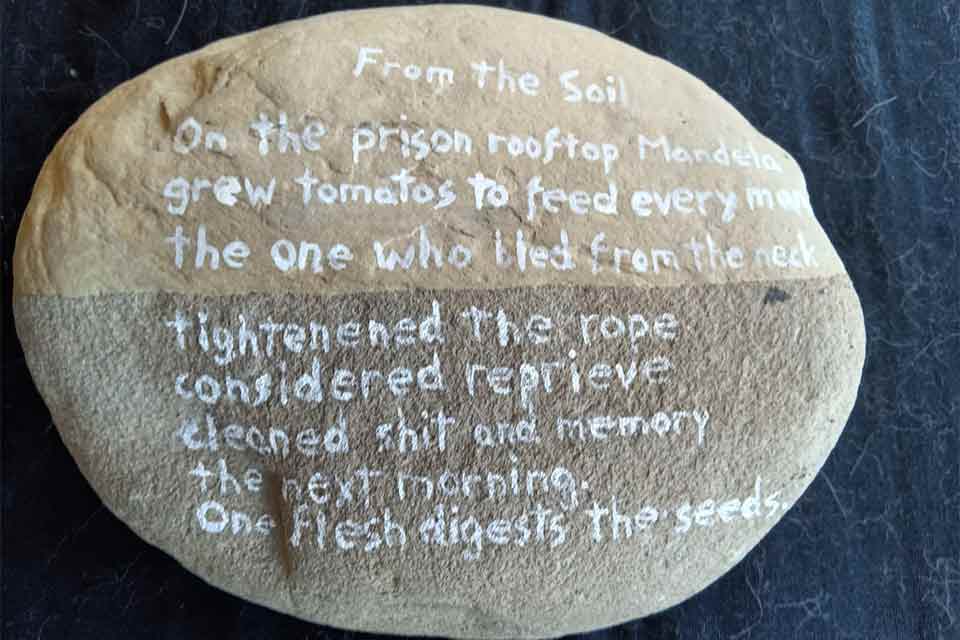 A poem written on a rock. Text reads: From the Soil  On the prison rooftop Mandela Grew tomatoes to feed every man The one who bled from the neck Tightened the rope Considered reprieve Cleaned shit and memory The next morning One flesh digests the seeds.