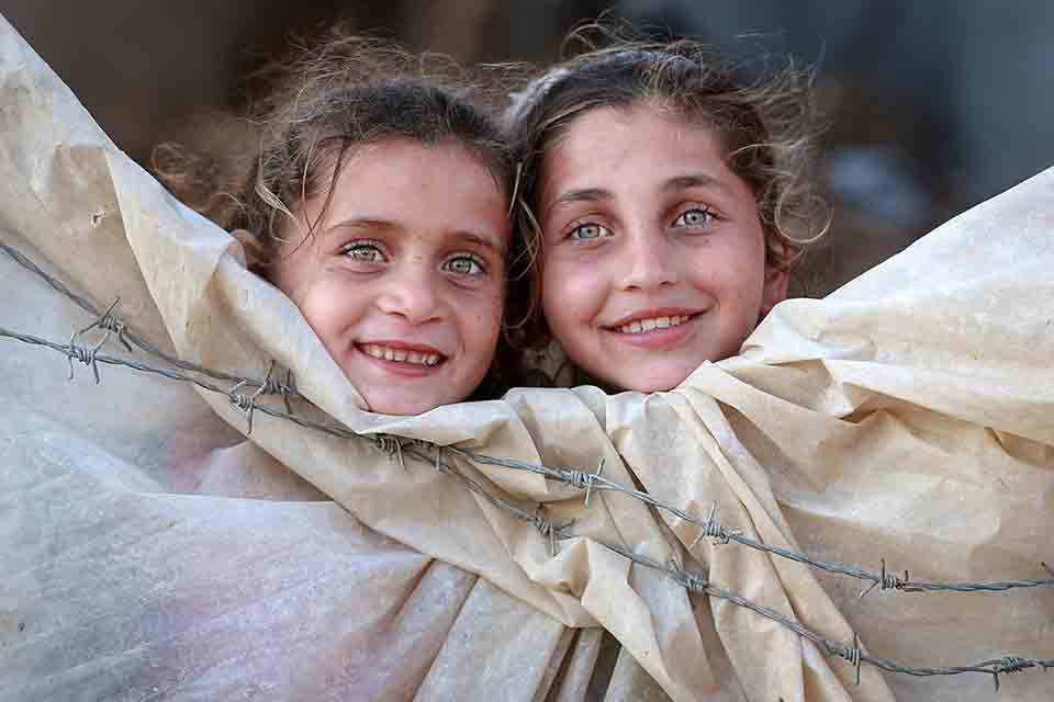 Two girls smiling. They are peering over canvas which protects them from the barbs on a wire fence