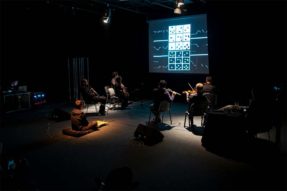 An ensemble of musician sit in puddles on light on a mostly darkened stage, watching a screen displaying information as they play stringed instruments