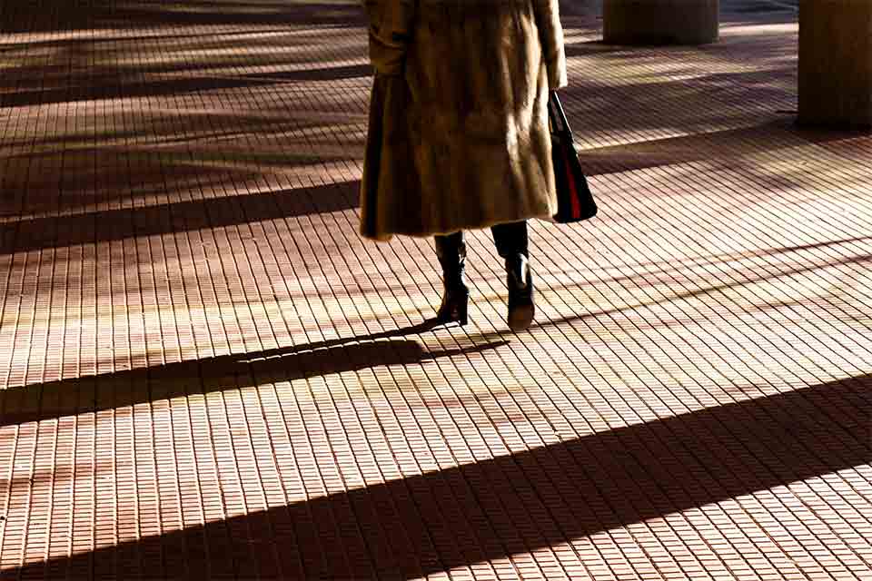 The bottom half of a woman, wearing a fur coat, standing near a crosswalk with long shadow crisscrossing the ground