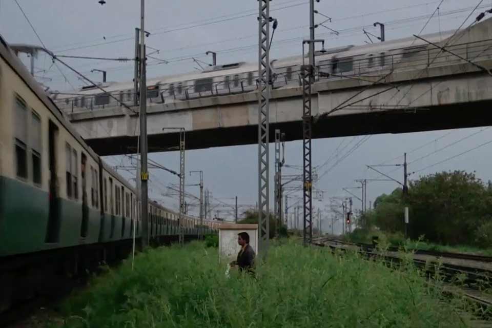 A man wades through tall grasses to approach a train in an industrial urban context