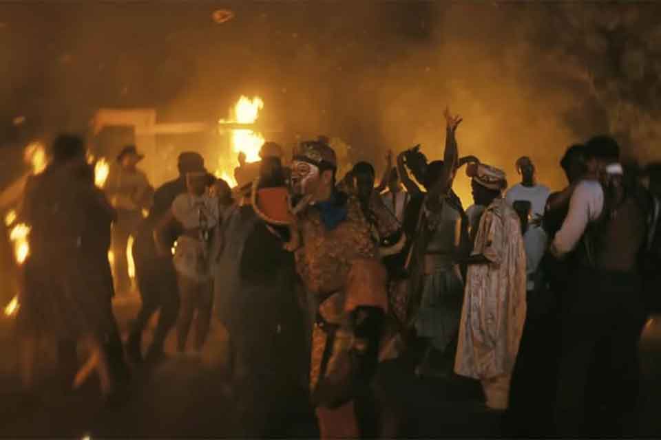 A group of people in various costumes dancing with flames rising from a structure in the background