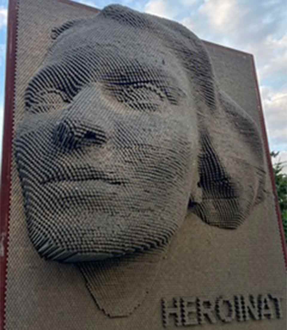A sculpture of a woman's face emerging from a flat wall. The text reads: Heroinat