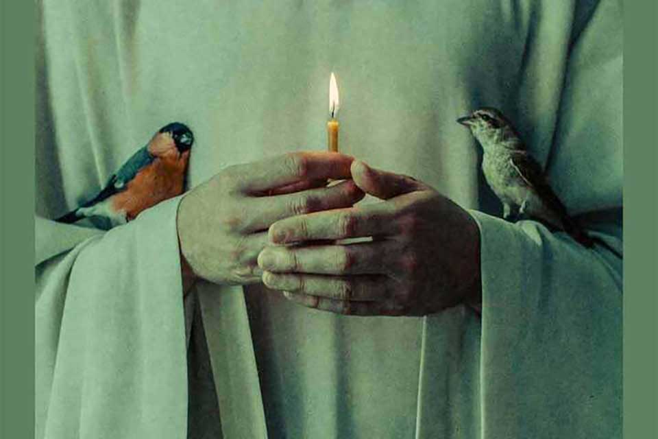 A pair of hands hold a candle. Each hand has a small bird perched on it.