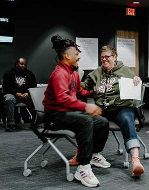 Two people sit in chairs in the center of a classroom. They are laughing