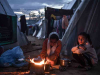 Three figures sit around a small fire outside of a canvas tent