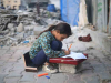 A young girl kneels in the rubble to use a displaced stone as a desk to work on schoolwork