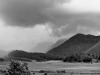 A moody black and white photo of a landscape at the foot of a tree covered mountain