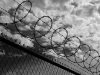 A black and white photograph looking up at the sky through a chain link fence topped with barbed wire