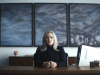 A woman sits at a desk in an office looking emotionlessly at the viewer