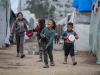 A group of refugee children carrying water basins, smiling as they walk together through a tent city