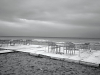 A black and white photos of empty tables and chairs on a patio overlooking a restless sea