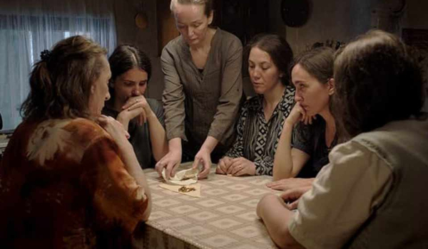 A group of women seated around a table. One woman, who is standing, appears to unfolding a cloth to reveal gold within