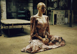 A bald woman sitting calmly in an ornate dress on a grubby floor