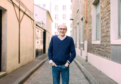 A photograph of a man standing with his hands in his front pants pockets looking at the viewer. He's standing on a narrow road between two buildings.