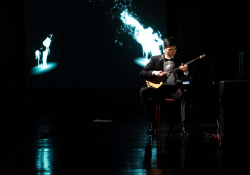 A man seated on a mostly darkened stage playing a saz