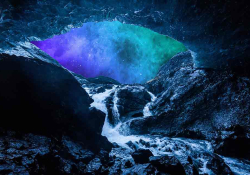 A river flows into the mouth of a darkened cave over rocks, creating a froth. The sky outside the cave is lit up with an aurora borealis
