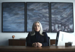 A woman sits at a desk in an office looking emotionlessly at the viewer