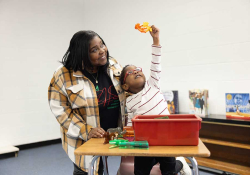 A woman stands beside a child as they look at a brightly colored toy in their hands