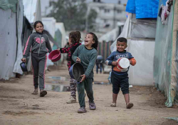 A group of refugee children carrying water basins, smiling as they walk together through a tent city