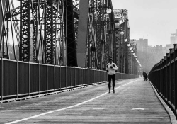 A black and white photograph of a person jogging toward the camera on a long suspension bridge