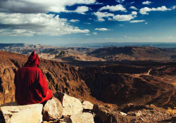 A photo of a figure, cloaked in red, sitting atop a mountain overlooking a wide vista below