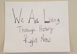 A hand-written sign. Text reads: We are living through history right now.