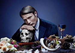 A promotional still of an actor, seated at a table, resting his chin atop his hand, which rests on a human skull