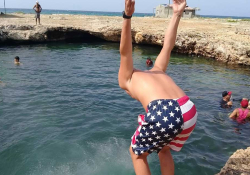 A figure clad in swimming shorts with an American flag on them faces away from the camera and is poised to jump into a natural swimming hole