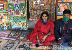 A woman clad in red sits with her husband (who is masked) on the ground. On the ground in front of them and mounted behind them on a wall of brightly colored paintings.