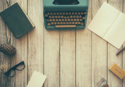 A typewriter, notepad, books, and glasses circle an empty spot on a rough woodgrain table