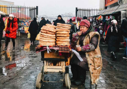 A bread vendor sits next to her piles of bread heaped on a bicycle on a stone plaza in winter