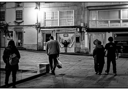 A black and white photo of a group of people, with its back to the viewer, walking toward a female figure, who stands in front of graffiti of a pair of angel's wings, painted on to the wall nearby