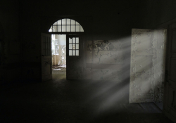 A photograph of a rundown room, mostly shrouded in shadow, with light streaking in an open door on the right