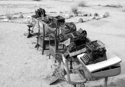 A black and white photograph of a line of typewriters on wooden desks on a beach. The desks and typewriters are covered with flotsam and sand.