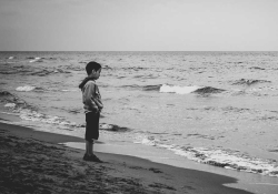 A somber black and white photograph of a boy standing on a beach looking at the ocean